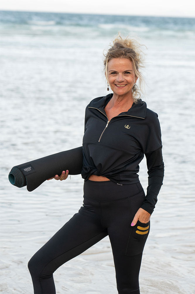 Ved havet med en yogamatte under armen. Genseren er knyttet i midjen over svarte tights.