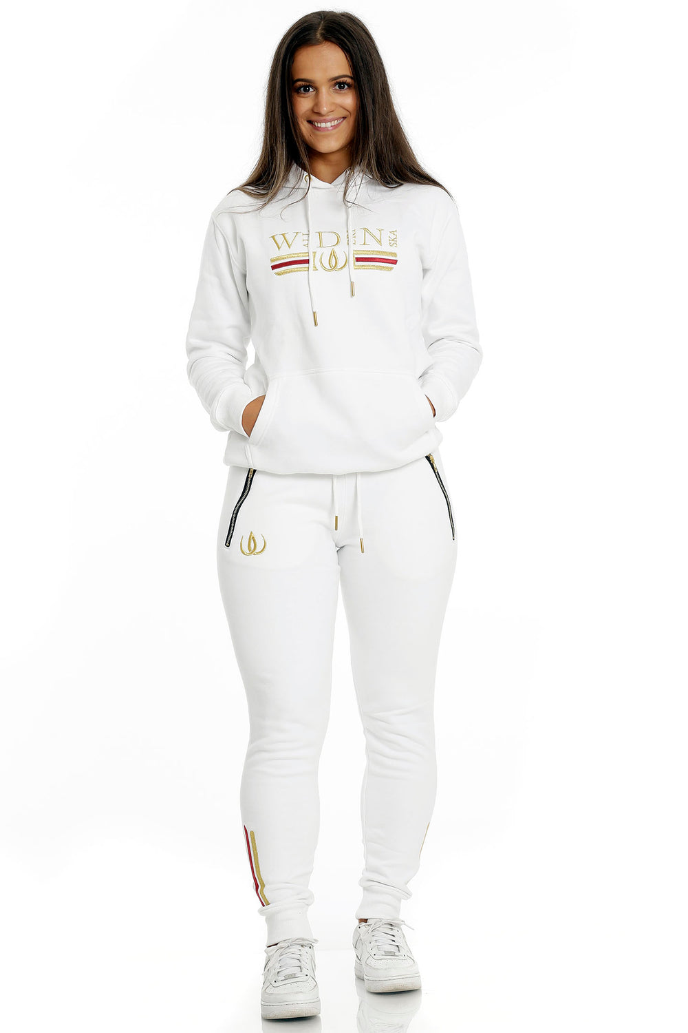 Stående i hvite joggers med gullogo og hvit hettegenser med gullbroderi, til hvite sneakers.