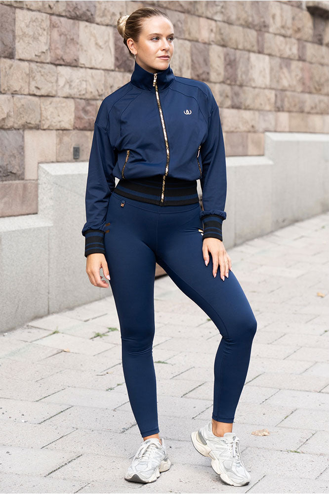 Utendørs i marineblå jakke med gullfargede glidelåser, marineblå tights og hvite sneakers.