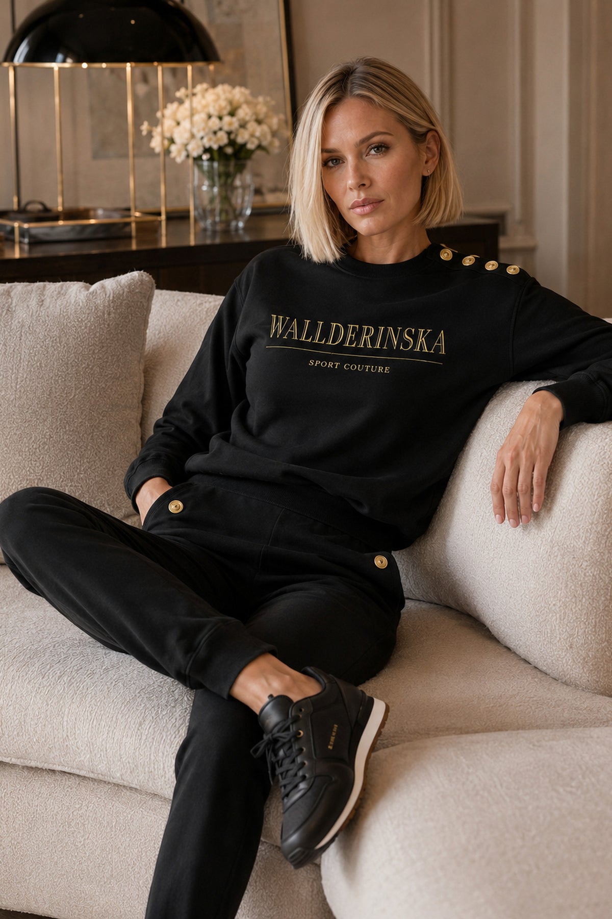 Edge Gold Button Sweater i svart med gulltekst på brystet og gullknapper på skulderen, båret i en lys sofa.