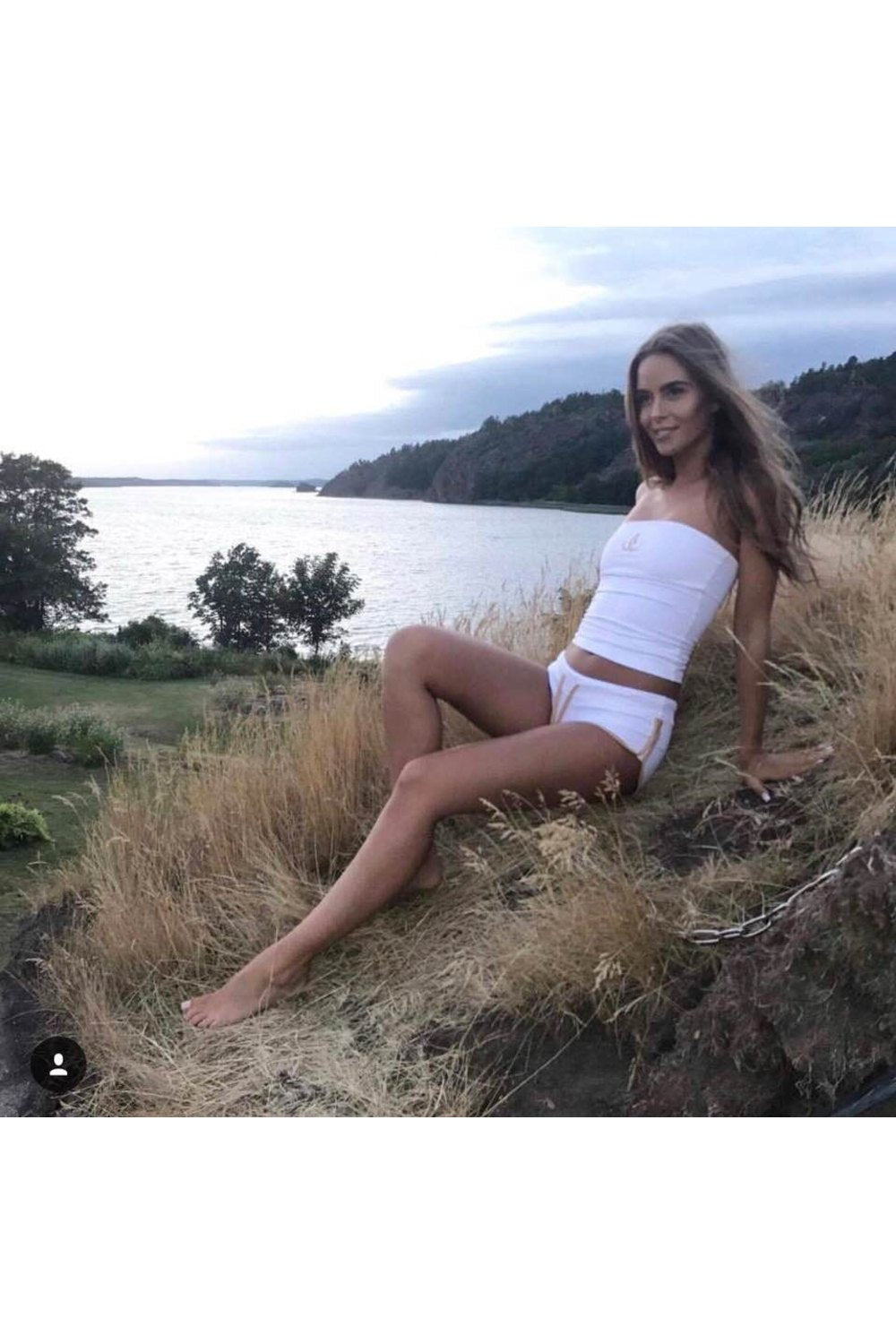 Kveldslys ved vannet. Modellen sitter i høyt gress i hvit bandeautopp og hvite shorts.
