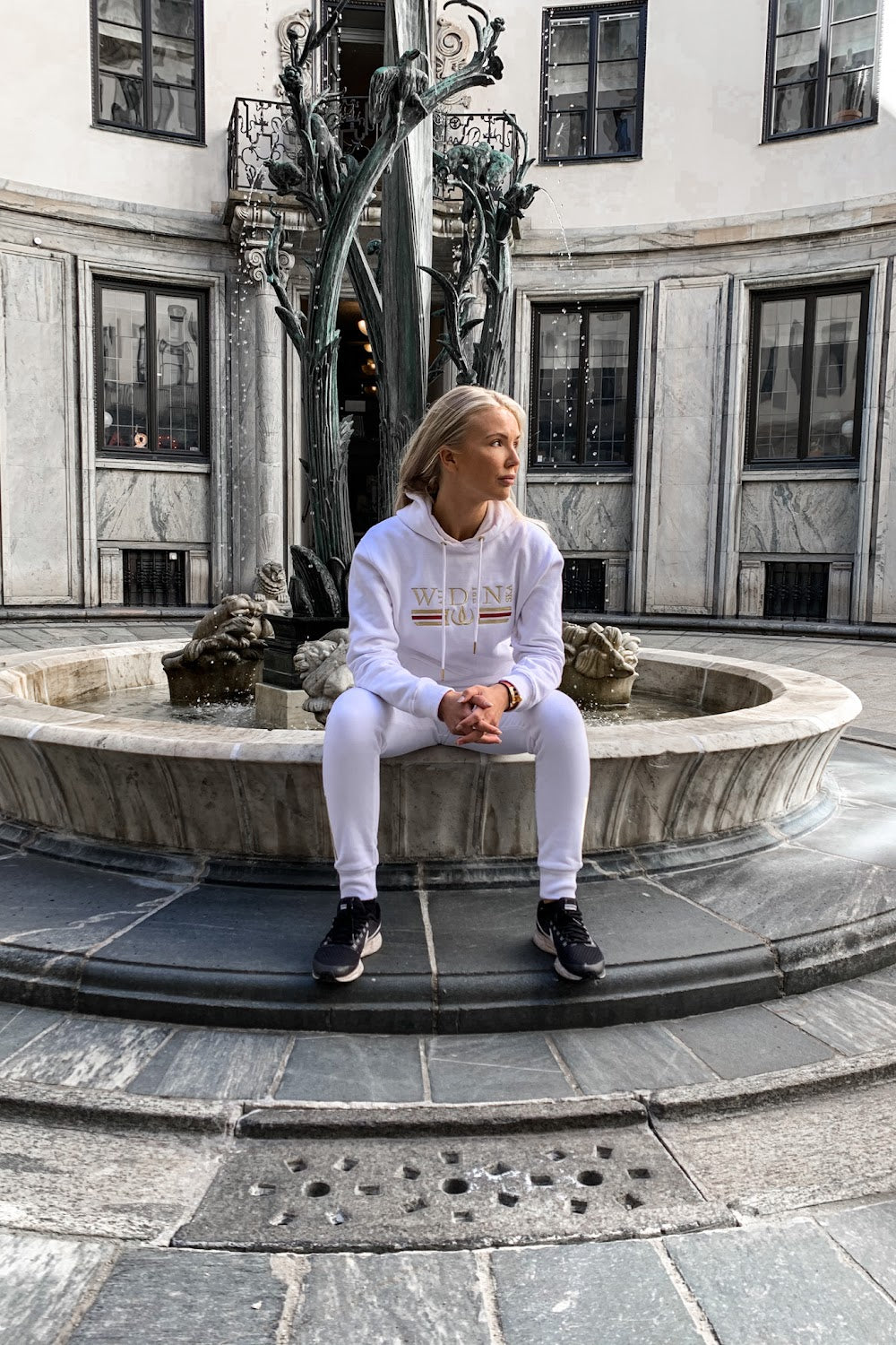 På fontenekanten sitter modellen i den hvite hettegenseren, med en statue i bakgrunnen.