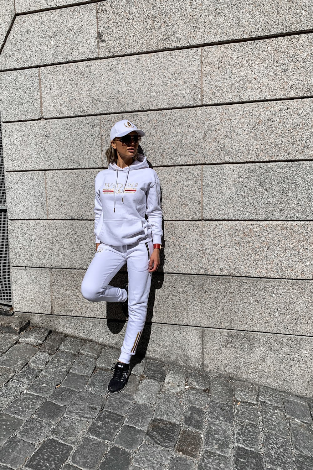 Mot steinveggen lener modellen seg med den ene foten trukket opp, i hvit hettegenser og hvite joggers.