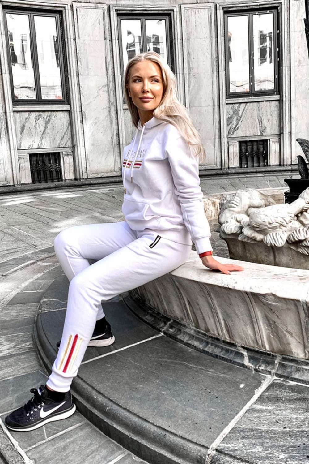 Modellen sitter ved en fontene i byen i den hvite hettegenseren og hvite joggers med gullstripe.