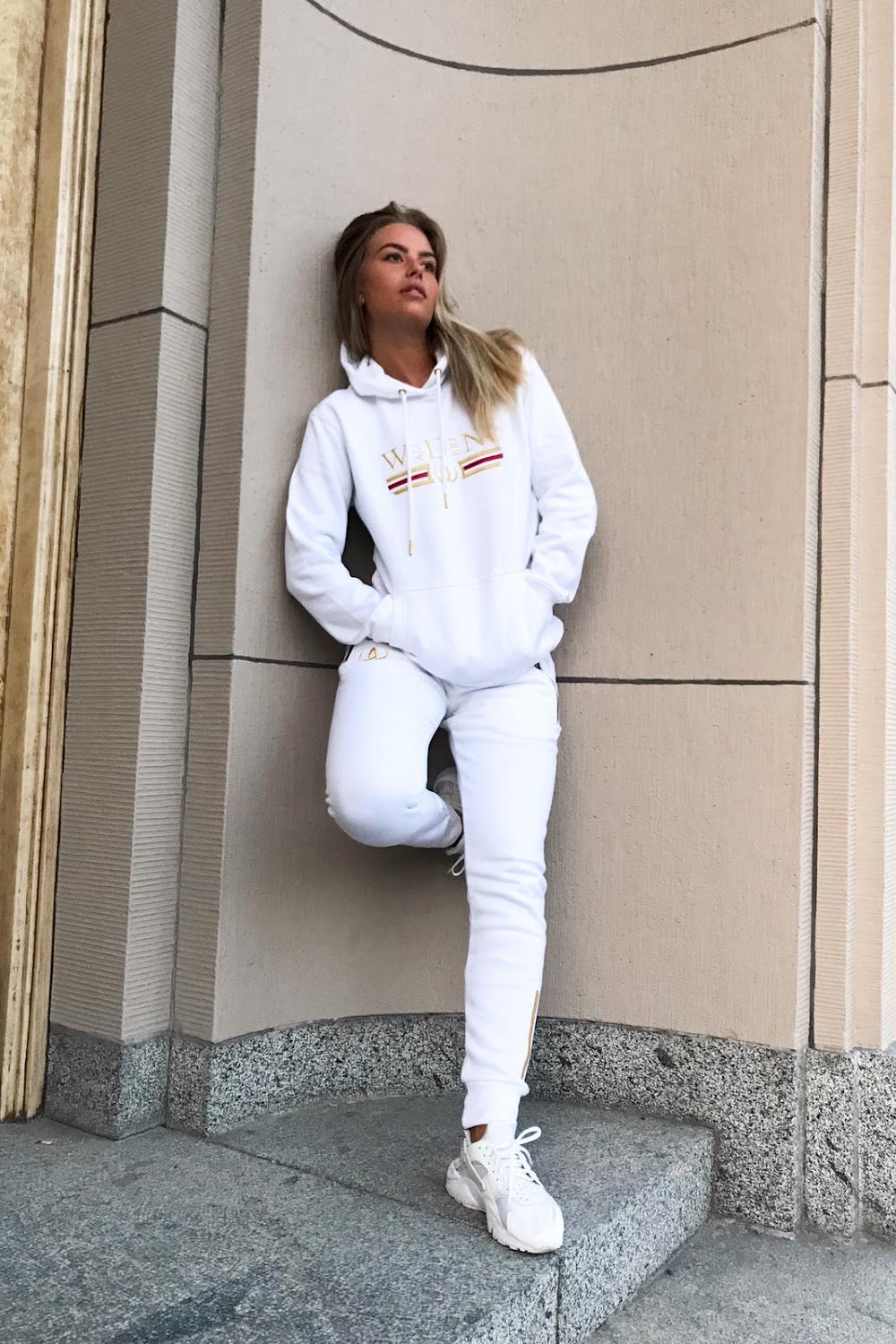 Mot husveggen står modellen i hvit hettegenser og hvite joggers, til hvite sneakers.