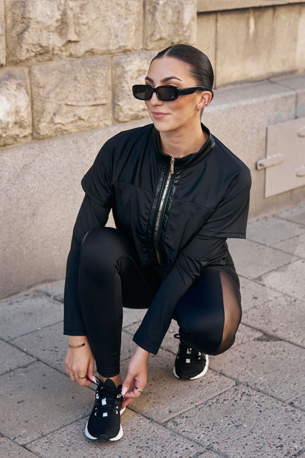 Ved fortauet knyter modellen skoen på huk, i den svarte jakken og svarte tights.