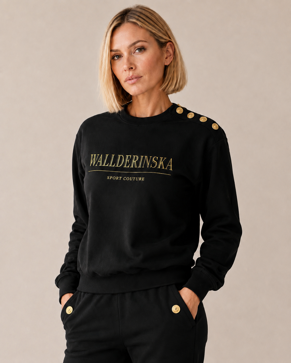 Modellen bærer Edge Gold Button Sweater i svart med gulltekst på brystet og gullknapper på den ene skulderen, til de svarte buksene med gullknapp ved lommen.