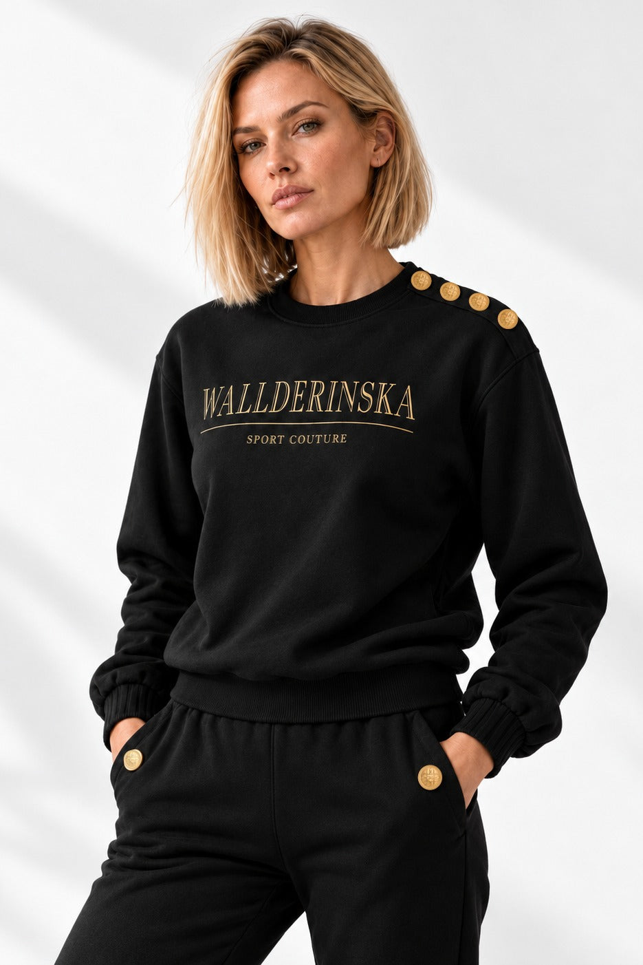 Edge Gold Button Sweater i svart forfra i lyst studio, med fire gullknapper på skulderen og gulltekst på brystet.